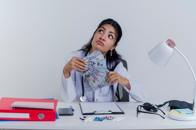 thoughtful-young-female-doctor-wearing-medical-robe-stethoscope-sitting-desk-with-medical-tools-holding-money-looking-side-isolated_141793-69054